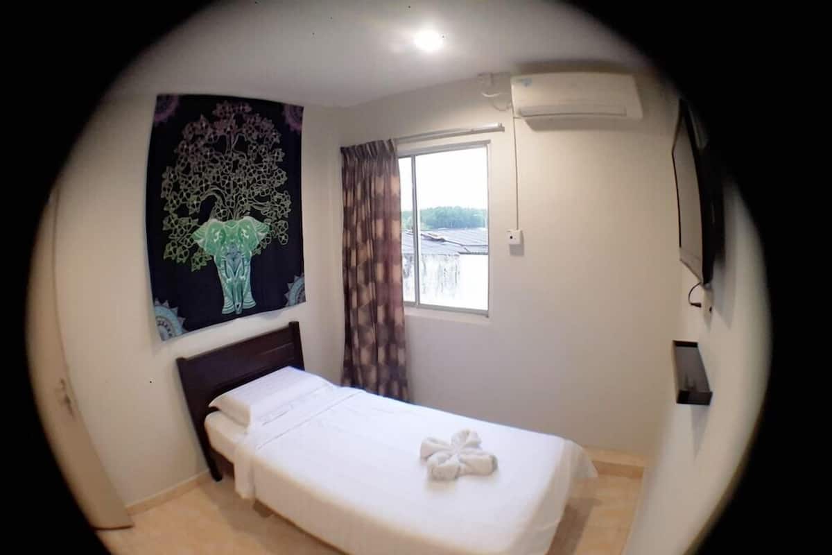 A single bed with a white bedspread and a towel, a window with curtains, an air conditioning unit, and a wall-mounted artwork.