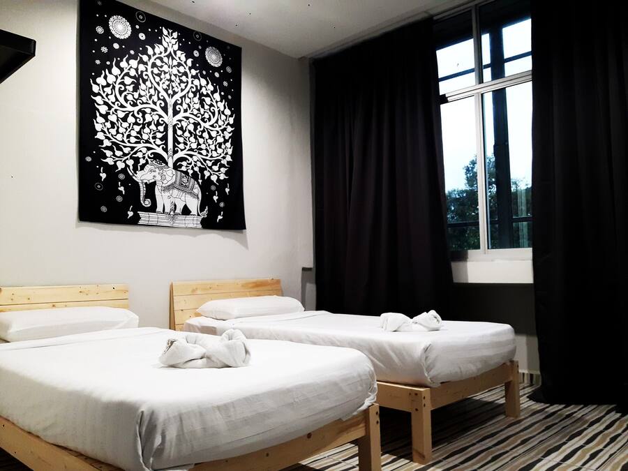 Habitación de hotel con tres camas individuales, un tapiz grande en blanco y negro, una ventana con cortinas negras y un suelo a rayas.