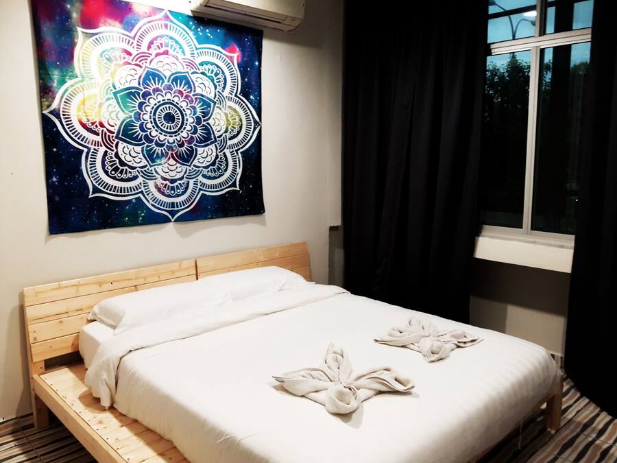 A hotel room with a wooden bed, white bedding, a large mandala artwork on the wall, and a window with curtains.