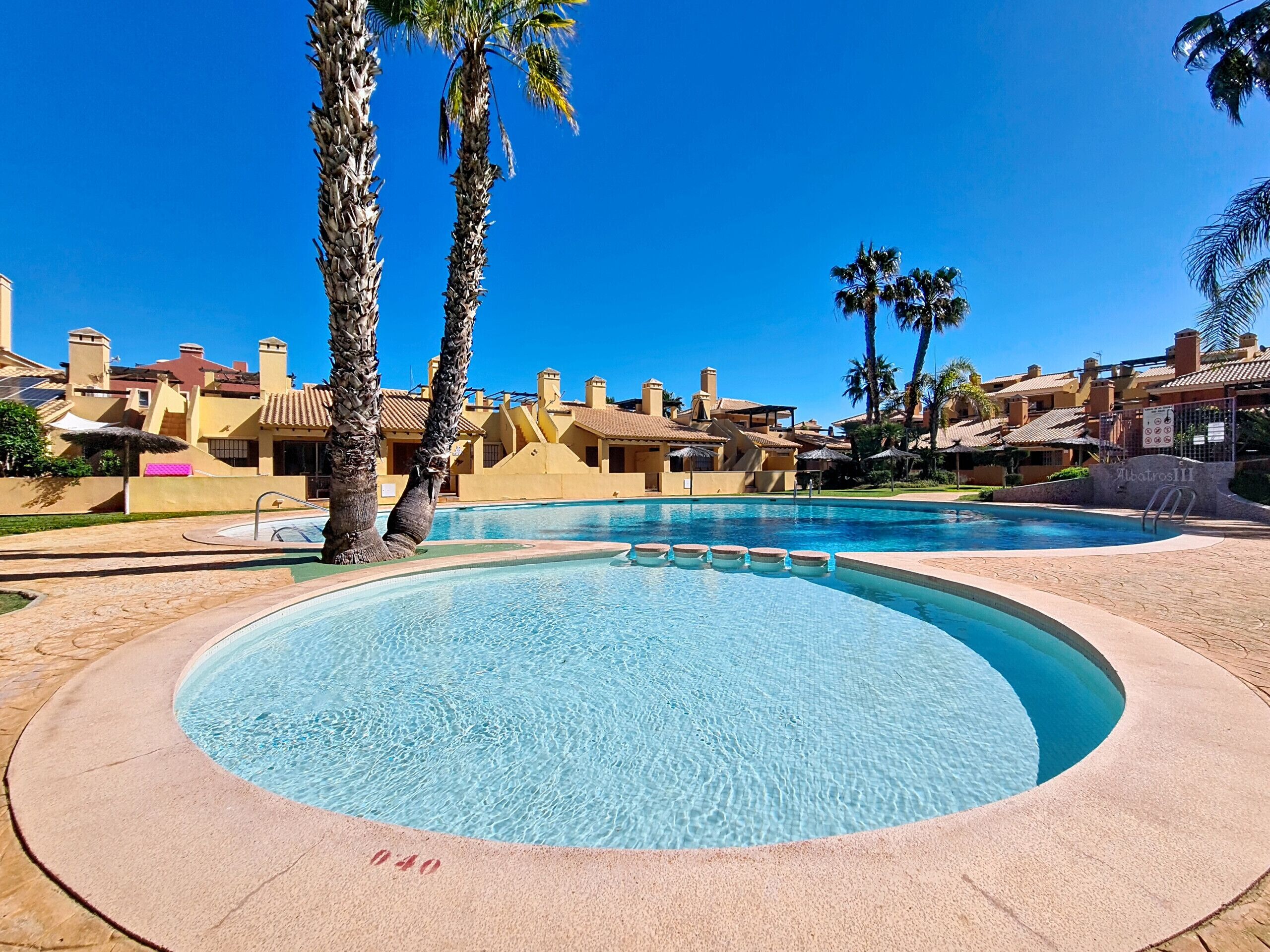 A pool area with clear blue water, palm trees, and buildings with tiled roofs.
