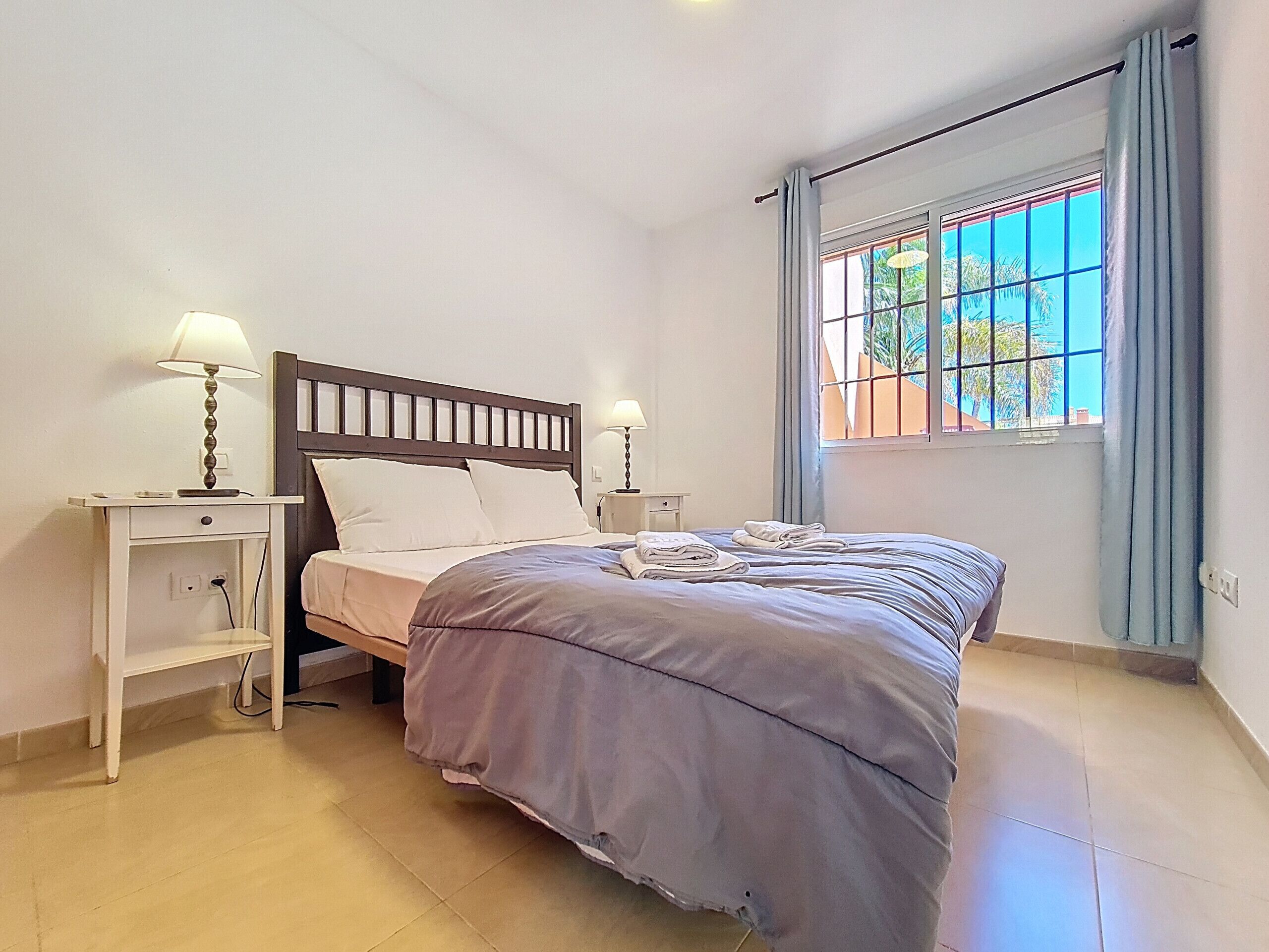 A bedroom with a bed, two bedside tables with lamps, a window with curtains, and a view of palm trees.