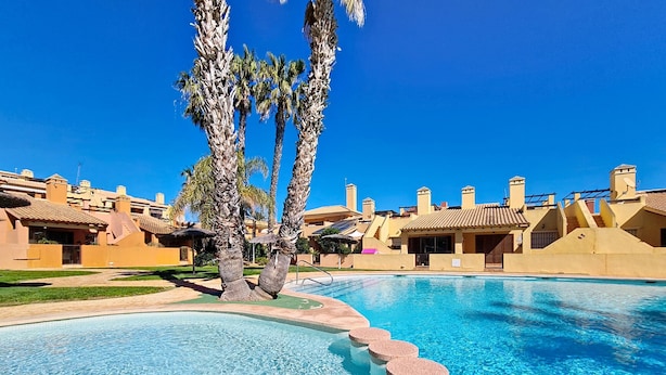 A swimming pool with a clear blue water feature, surrounded by palm trees and resort-style buildings.