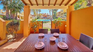 A wooden table with plates and glasses, surrounded by wicker chairs, under a pergola with a view of a swimming pool and palm trees.