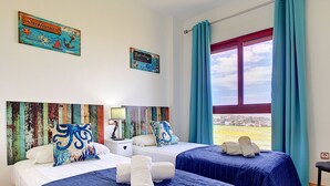 A hotel room with two beds, each with blue bedding and decorative pillows, a nightstand with a lamp, and a view of the sea through a window with blue curtains.