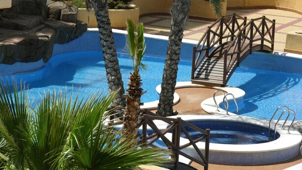 A swimming pool with a wooden bridge, palm trees, and a hot tub.