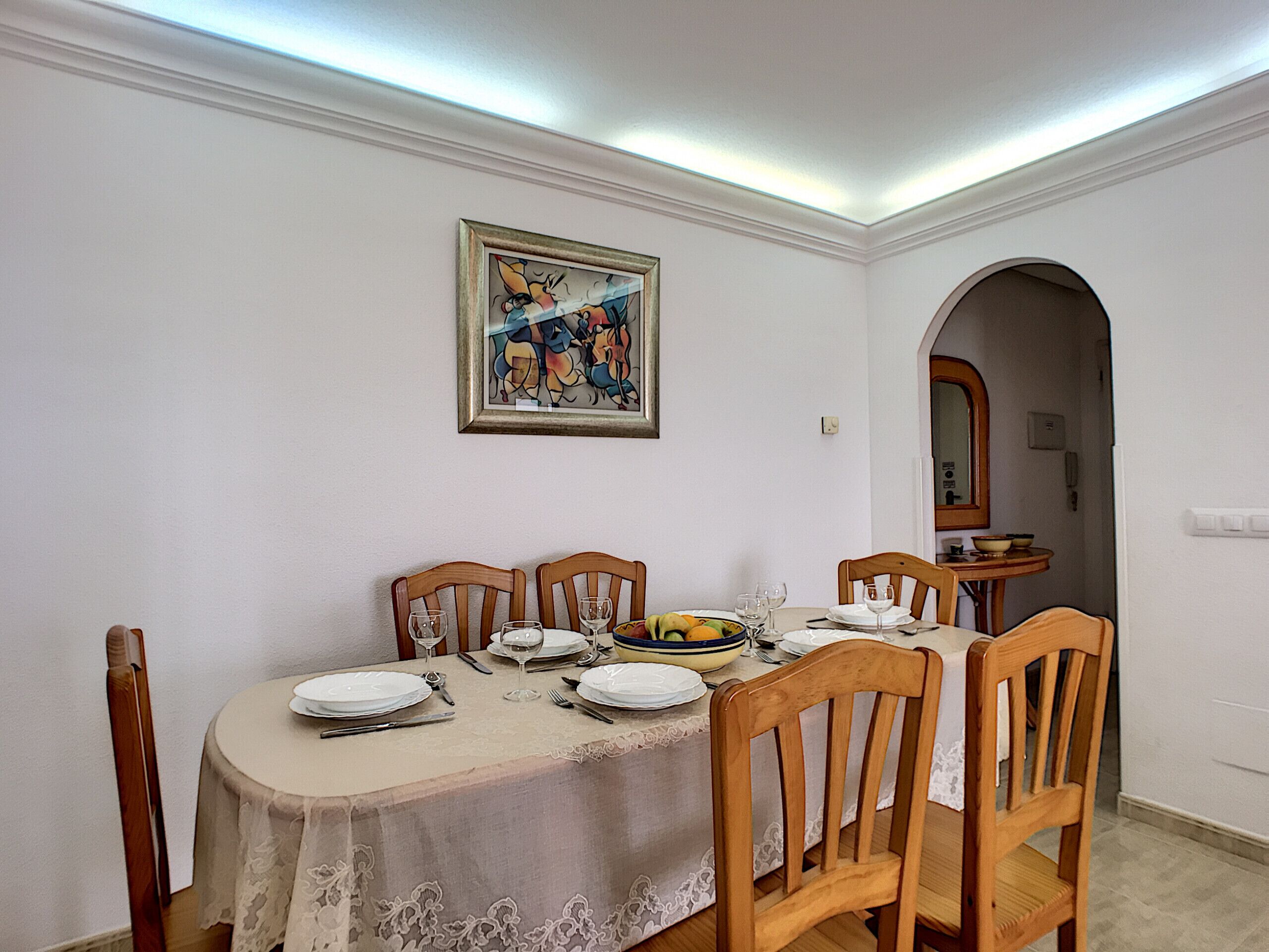 Comedor con una mesa dispuesta para cuatro, paredes blancas y un cuadro enmarcado sobre la mesa.