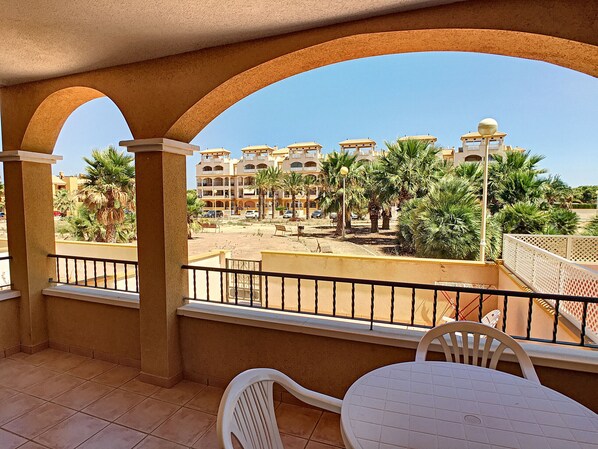 A balcony with a view of a courtyard, palm trees, and a building with multiple balconies.