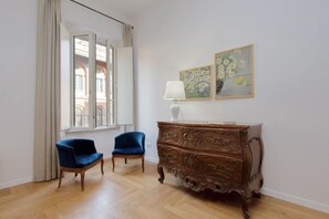 A room with a wooden dresser, two chairs, a window with curtains, and two framed floral paintings on the wall.