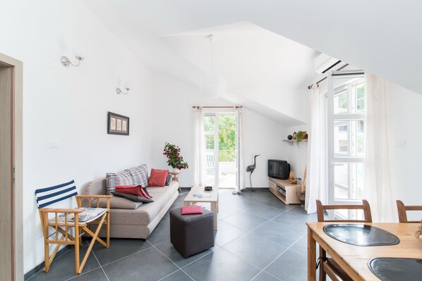 Un salon moderne comprenant un canapé, une chaise en bois, une petite table et une table à manger. Une fenêtre avec des rideaux offre une vue sur la verdure à l’extérieur.