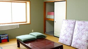 A room with tatami flooring, a low wooden bed with pillows, a wardrobe with shelves, and a large window. - Goishigahama House (Shimoda)