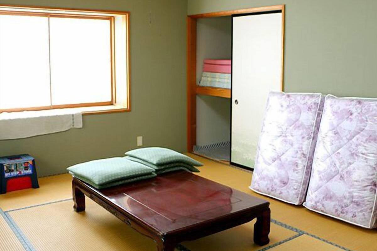 Une pièce avec un sol recouvert de tatamis, un lit bas en bois avec des oreillers, une armoire avec des étagères et une grande fenêtre.