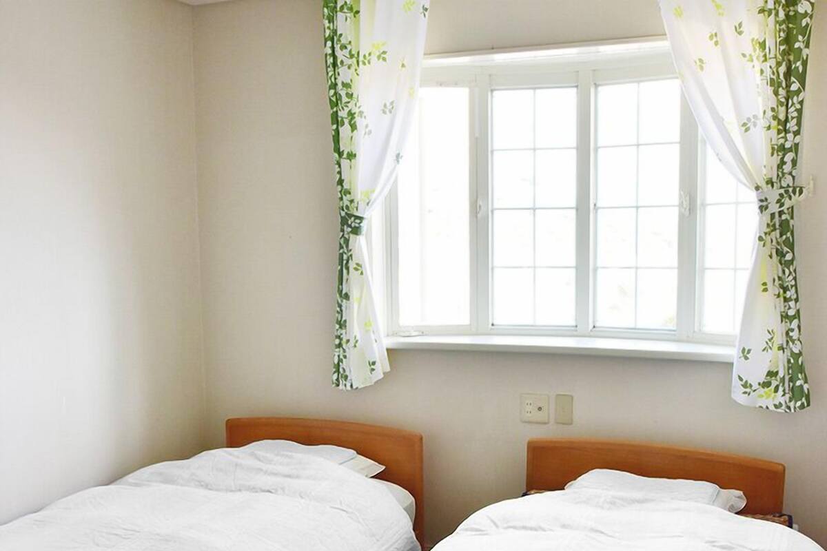 Une chambre avec deux lits simples, une fenêtre avec des rideaux blancs et un mur uni.