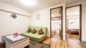 A room with a green sofa, a small table with a vase of flowers, a clock on the wall, and a door leading to another room with a bed. - Minhouses (Kyoto)