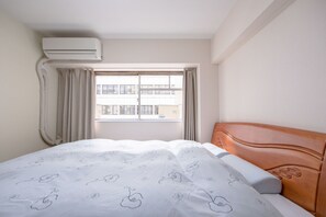 A hotel room with a bed, an air conditioning unit, a window with curtains, and a view of buildings outside. - Minhouses (Kyoto)