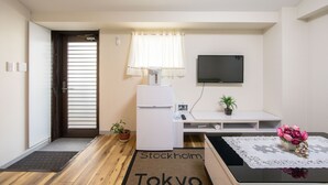 A compact living space with a kitchenette, a flat-screen TV mounted on the wall, a small potted plant, and a decorative vase with flowers on a table. - Minhouses (Kyoto)