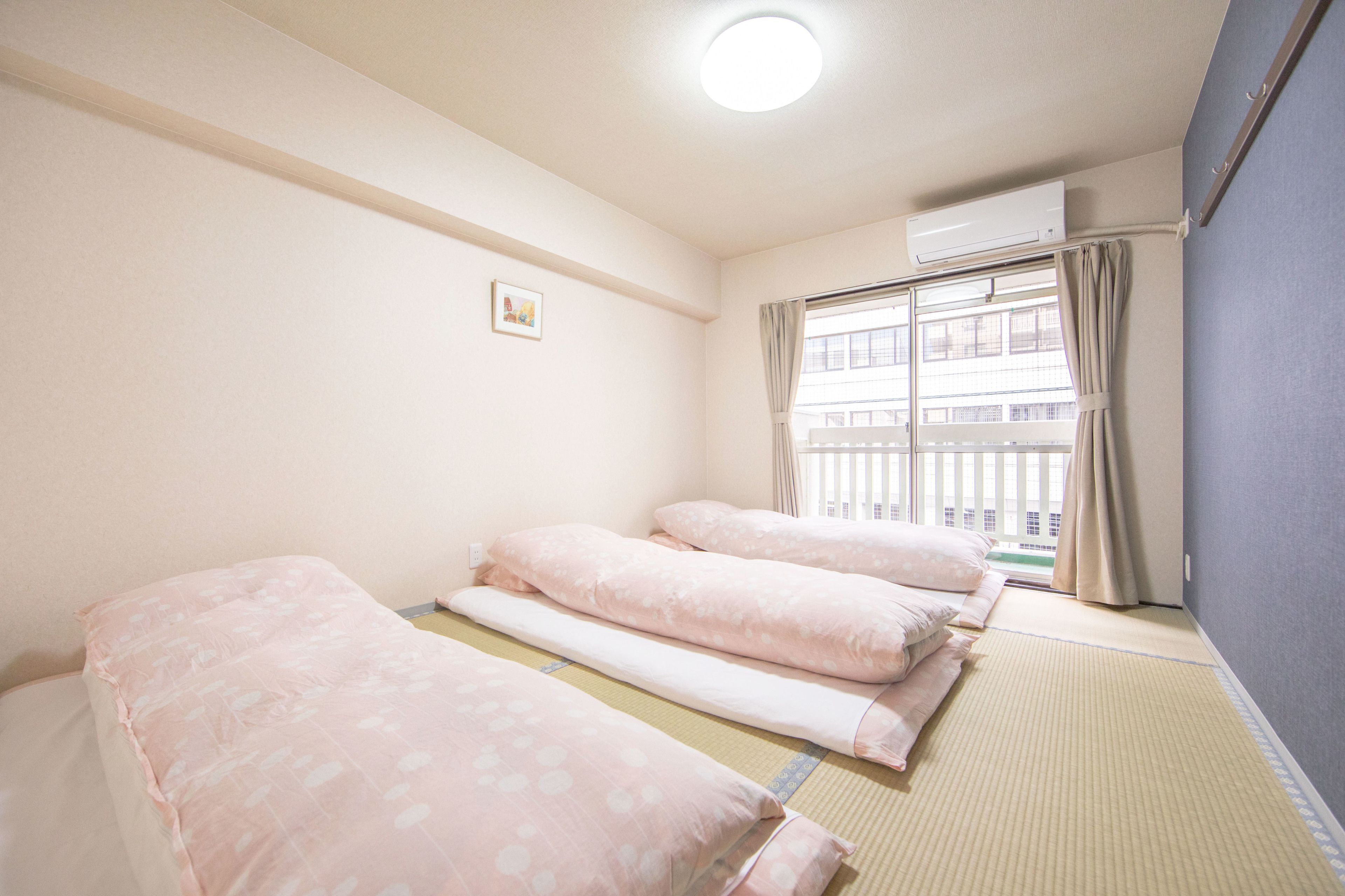 A room with three beds arranged in a row, a window with curtains, and an air conditioning unit on the wall.