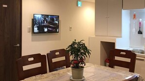 A dining area with a table covered in a floral tablecloth, surrounded by wooden chairs. A potted plant is on the table, and a television is mounted on the wall. - Minhouses (Kyoto)