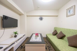 A modern living room with a flat-screen TV, a small table with a floral centerpiece, a green sofa with brown cushions, and a wall-mounted clock.