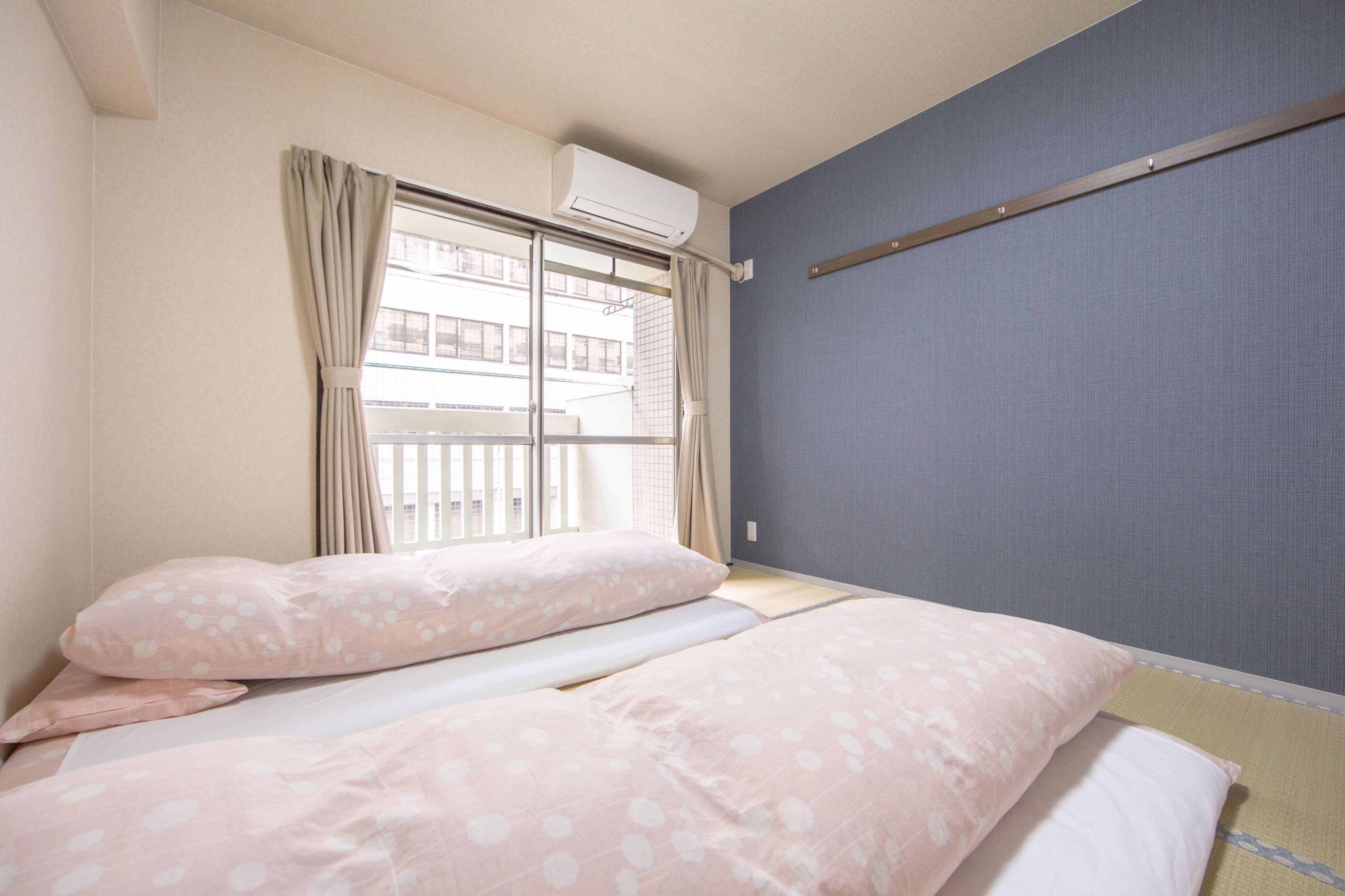 A hotel room with a bed, curtains, an air conditioning unit, and a view of buildings outside.