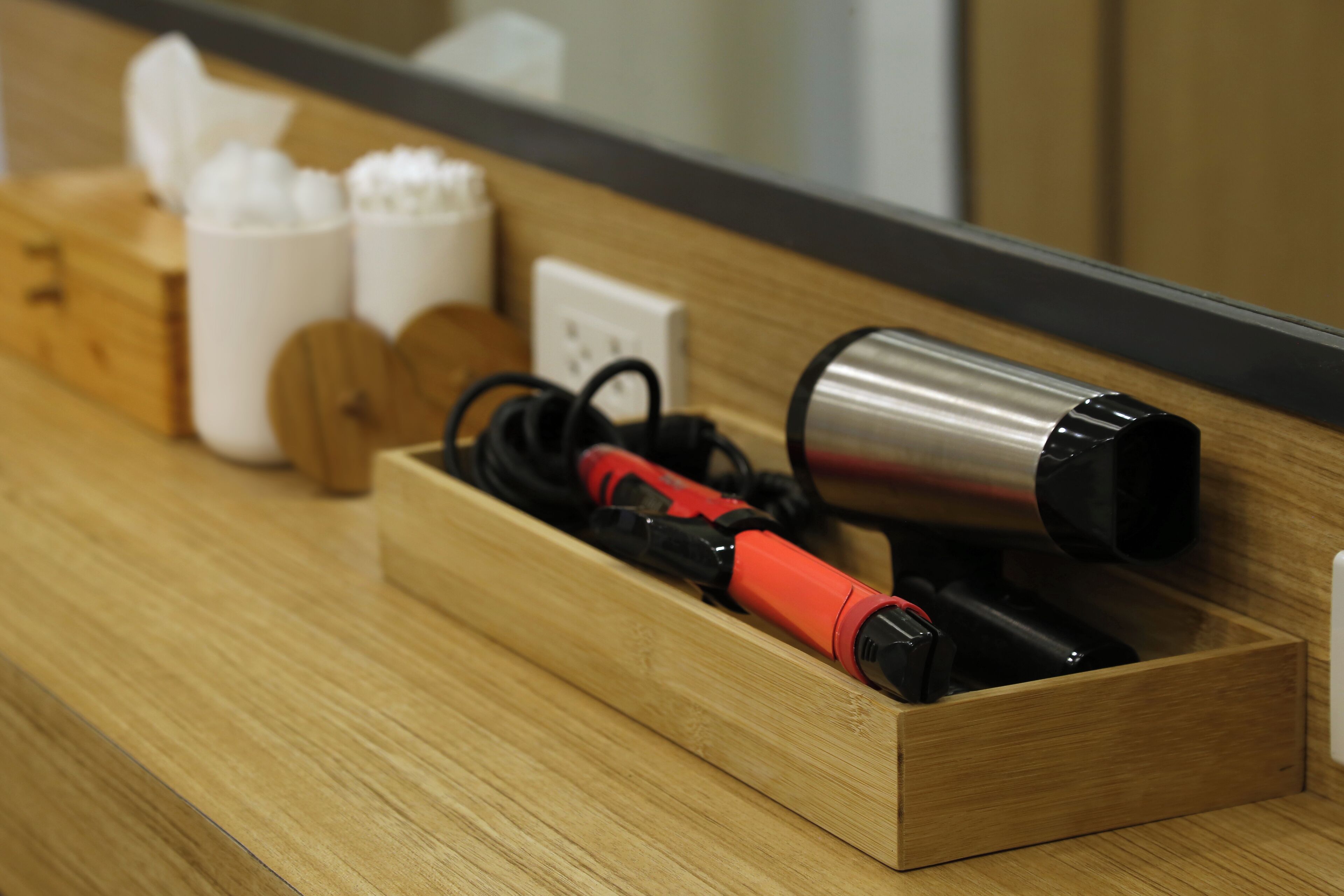 Un tiroir en bois contenant un sèche-cheveux rouge et noir et un objet cylindrique argenté, qui pourrait être un radiateur portatif, est posé sur une surface en bois. Un miroir reflète une partie de la pièce.