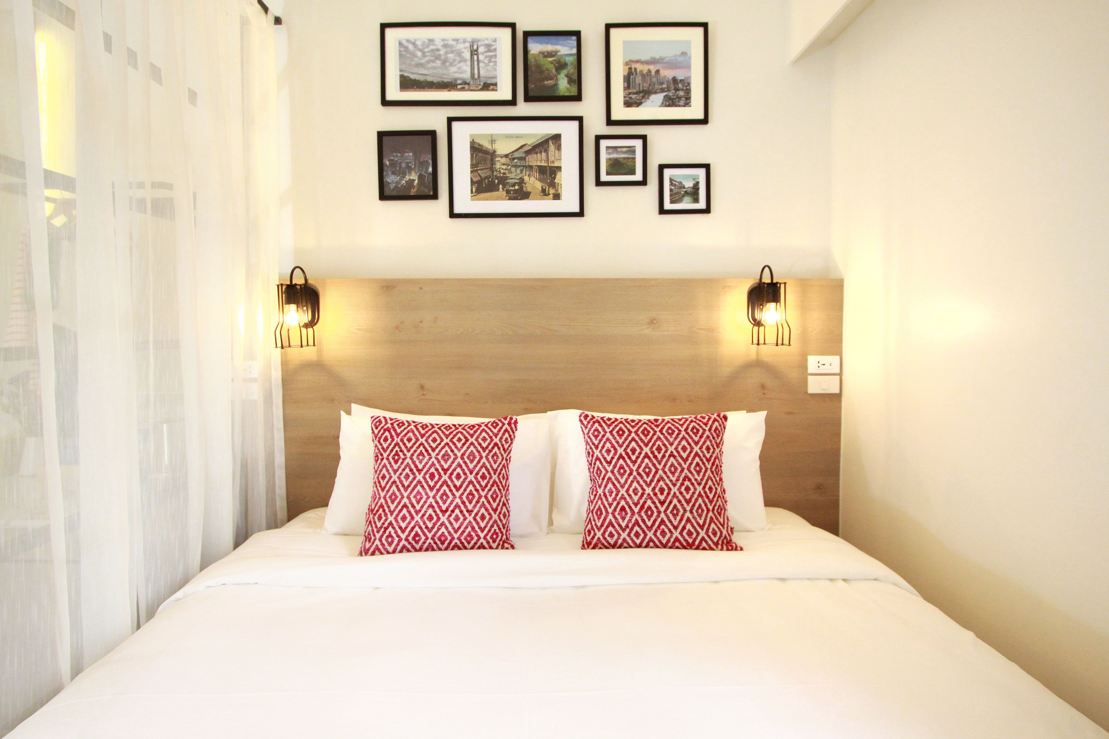 Camera d'albergo con una testiera in legno, due cuscini rossi con motivo decorativo, una coperta bianca e diverse immagini incorniciate appese al muro.