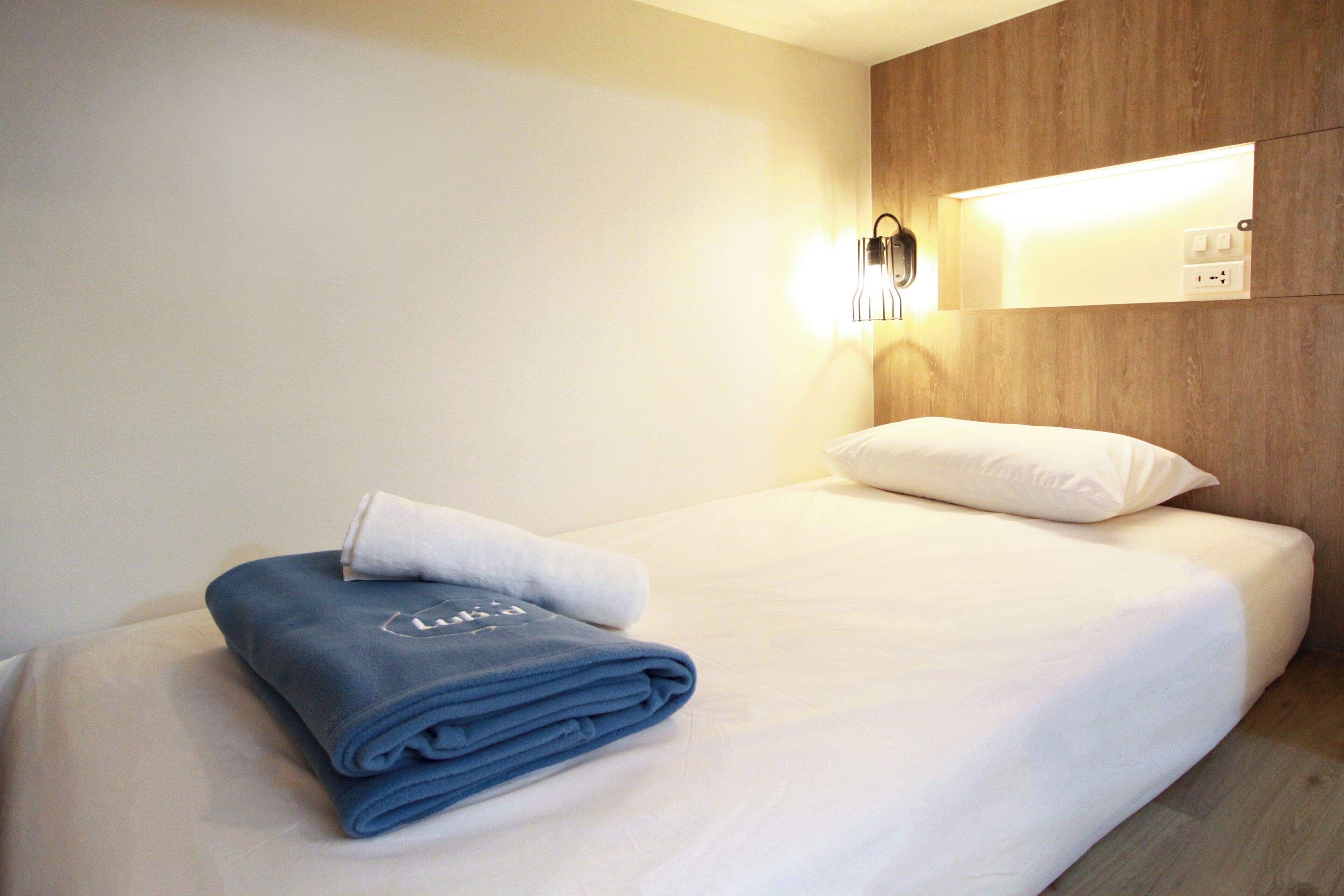 A neatly made bed with a folded towel and a pillow, a wall-mounted light, and a wooden panel.