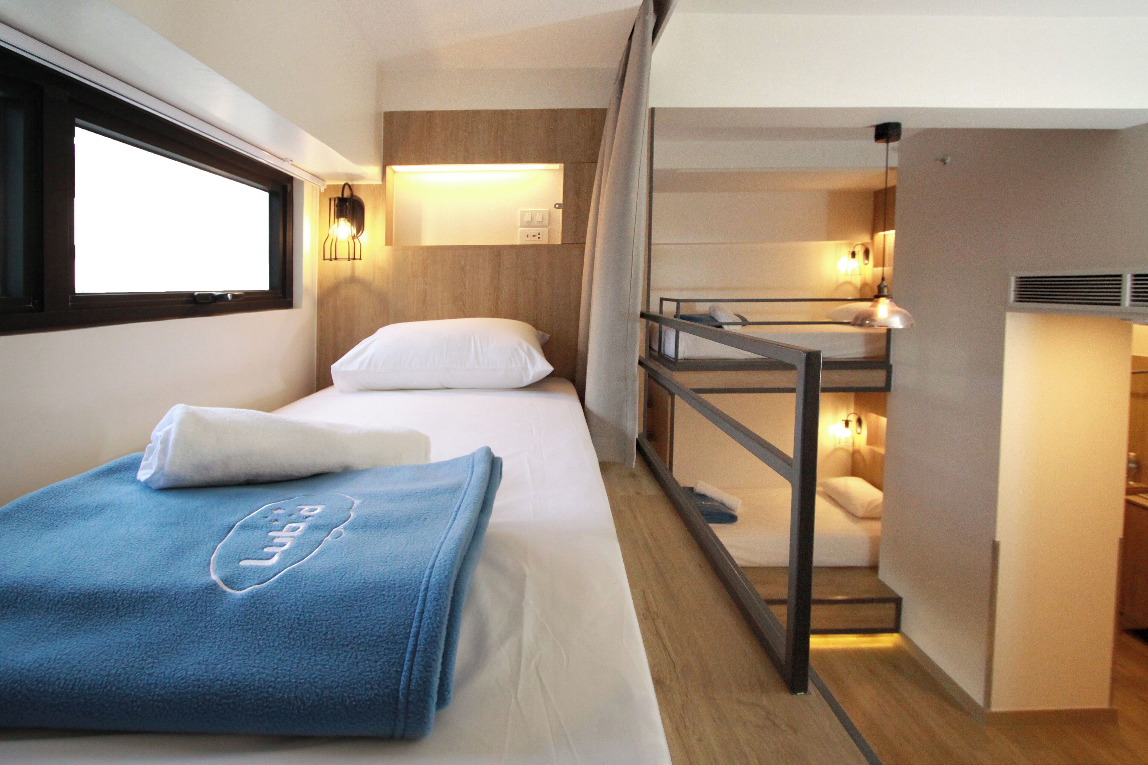A hotel room with a single bed, a bunk bed, a window with blinds, and a wall-mounted light fixture.