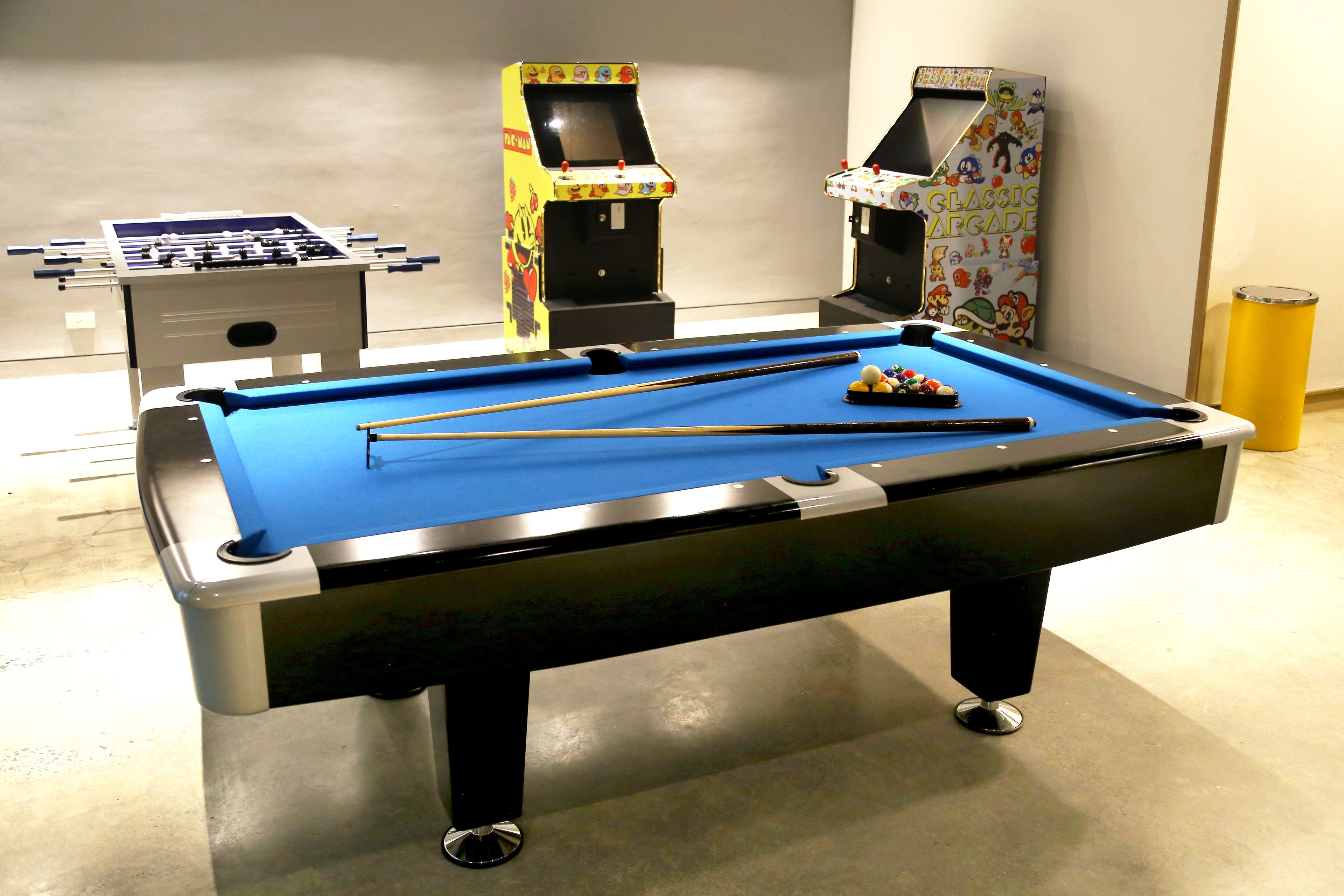 A pool table with a blue felt surface, two foosball tables, and a trash can.