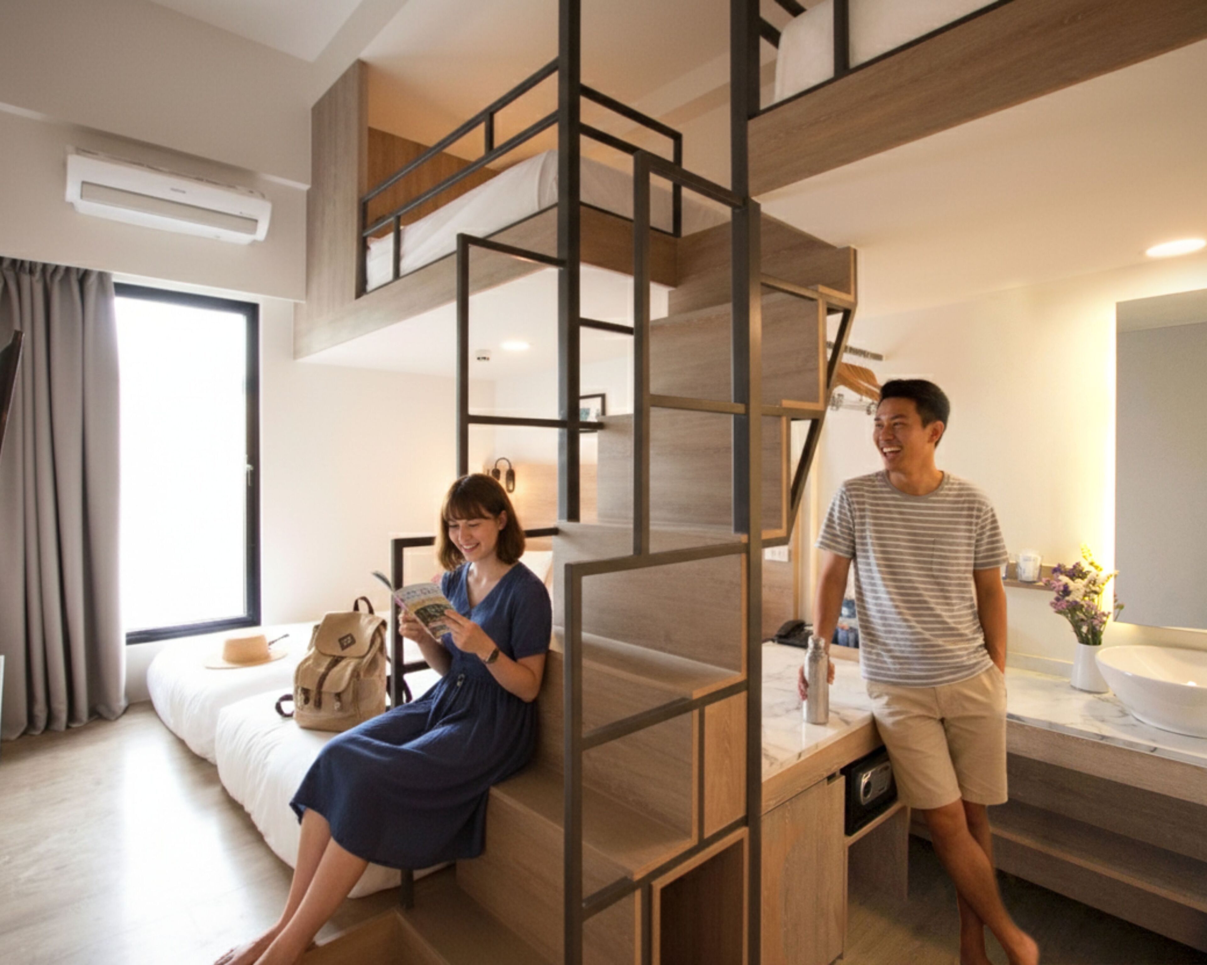 A modern hotel room with a bunk bed, a large window with curtains, a bedside table with a lamp, and a bathroom with a sink and mirror.