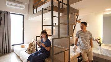 A modern hotel room with a bunk bed, a large window with curtains, a bedside table with a lamp, and a bathroom with a sink and mirror.