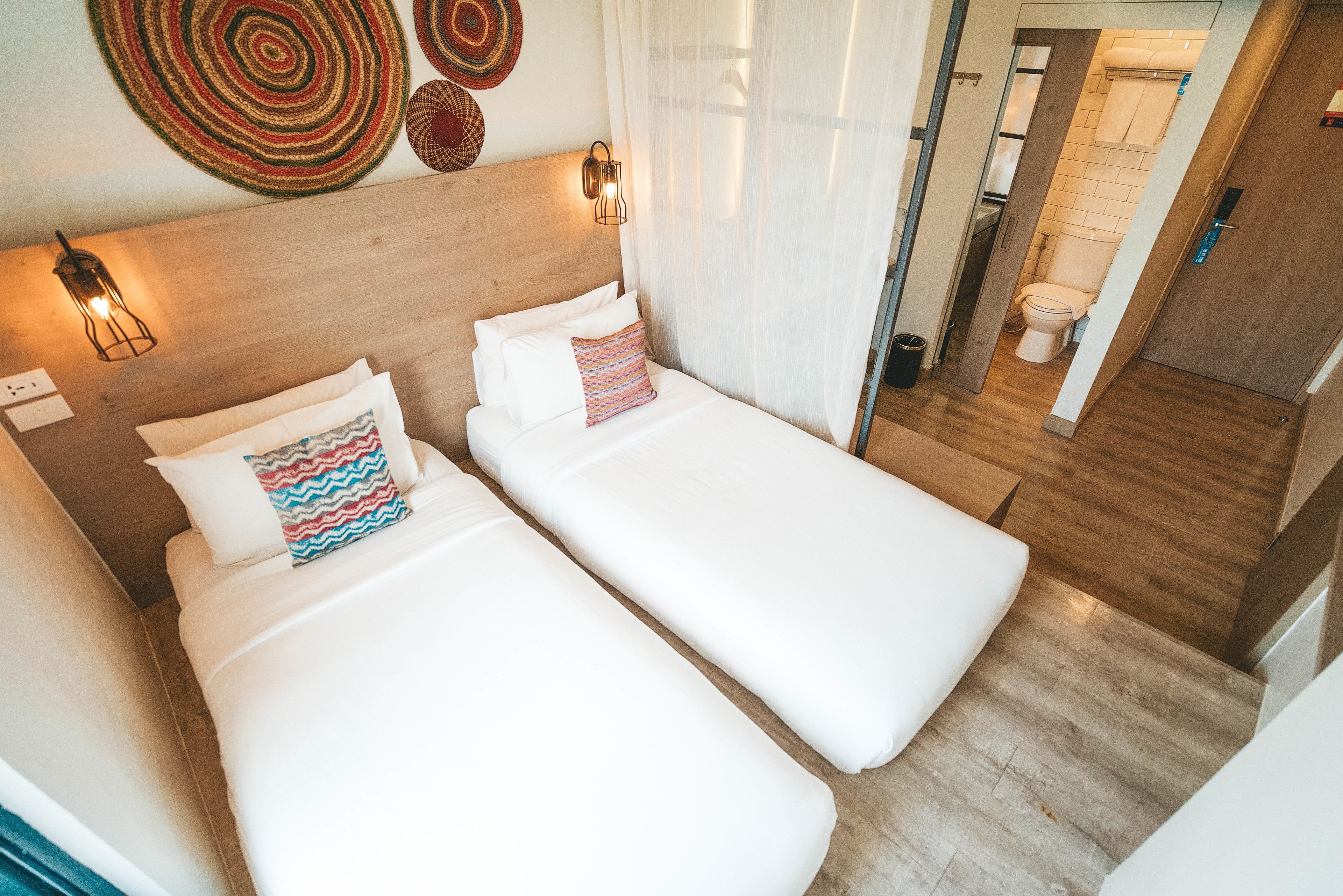 A hotel room with two single beds, a wooden headboard, a wall-mounted light fixture, and a view of the bathroom.