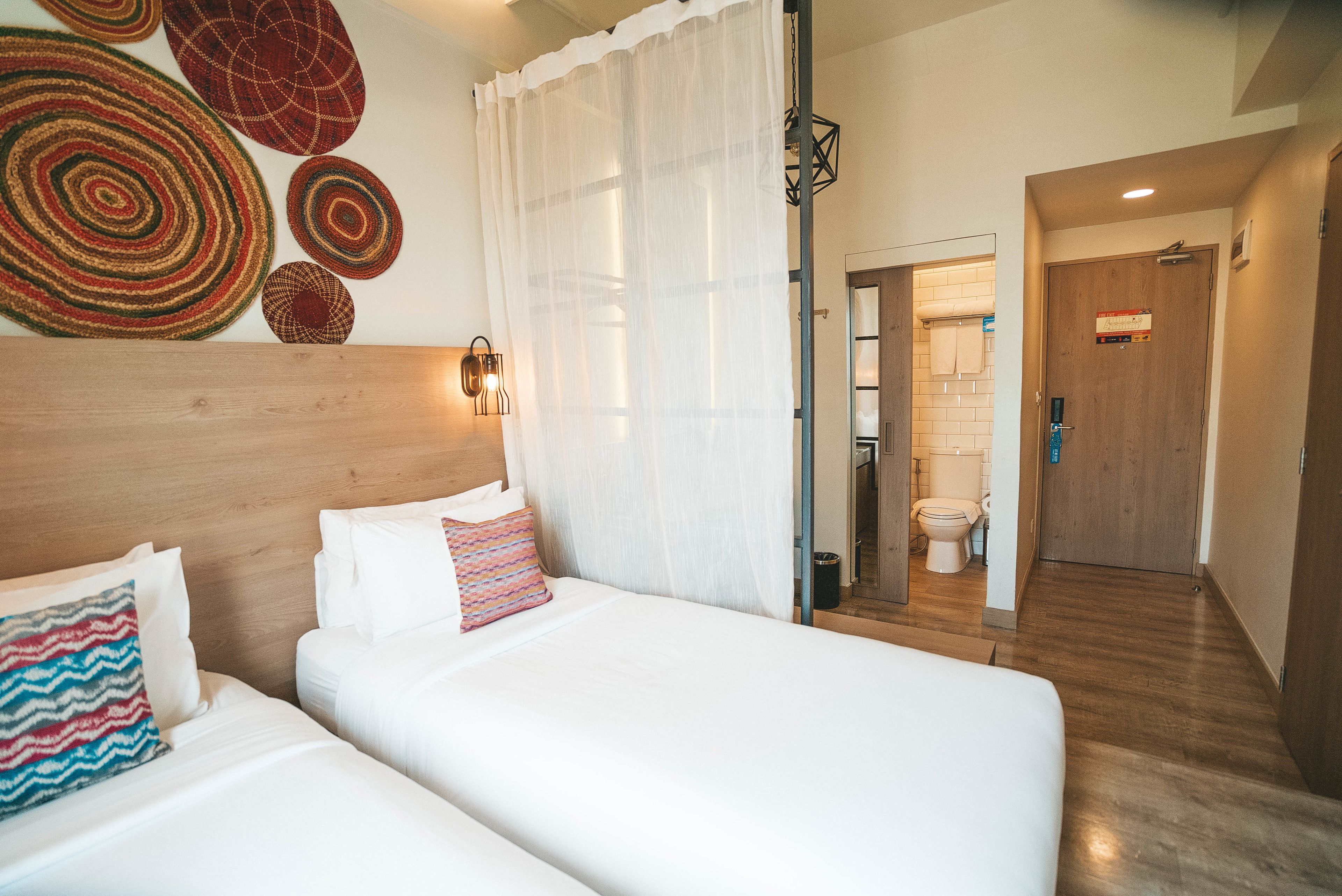 Camera d'albergo con due letti, una testiera in legno, una parete decorata con tappeti circolari e un bagno visibile attraverso una porta aperta.