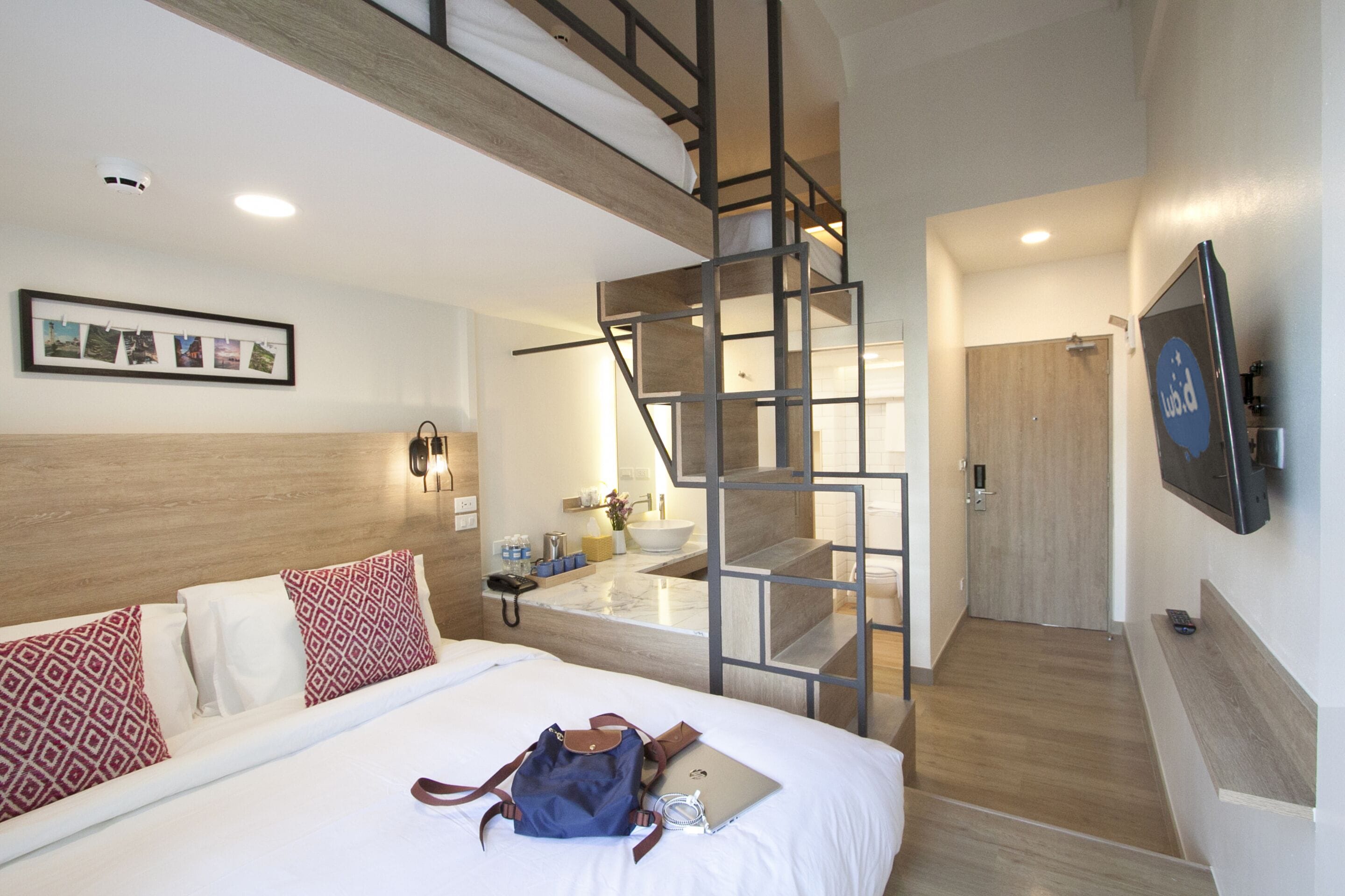 Una habitación de hotel con una cama, un escritorio, una computadora portátil, una mochila, una escalera y un televisor.