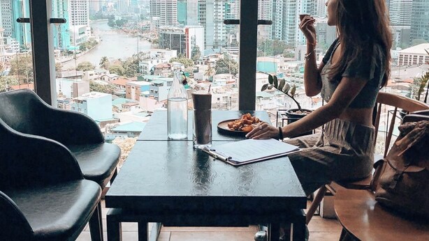 A woman dining at a table with a city view, a glass of drink, and a plate of food.