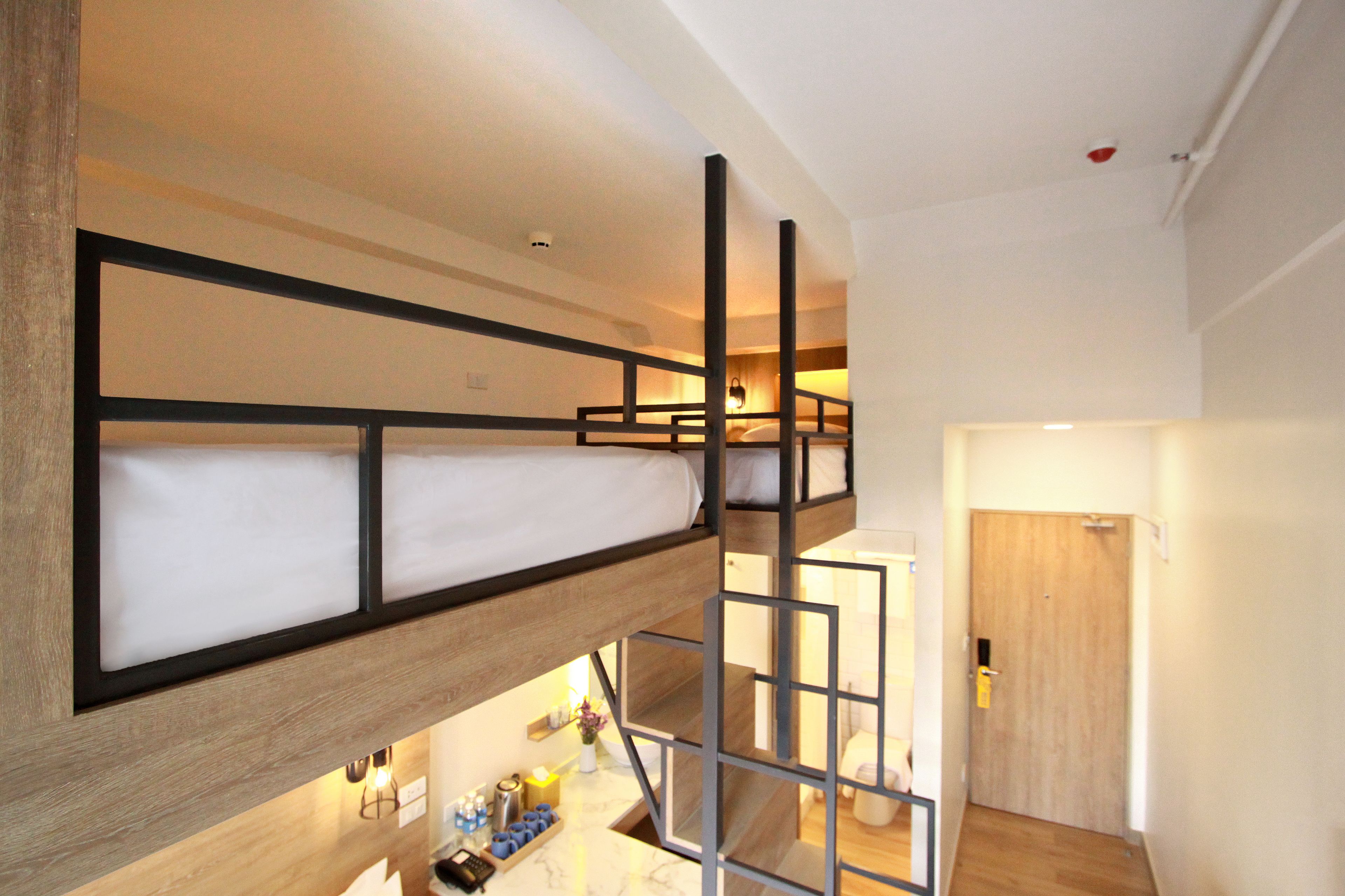 Una cama litera con estructura de madera y colchón blanco. Hay una mesita con un florero y velas. Se ve una puerta de madera al fondo.