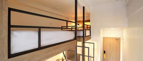 Una cama litera con estructura de madera y colchón blanco. Hay una mesita con un florero y velas. Se ve una puerta de madera al fondo.