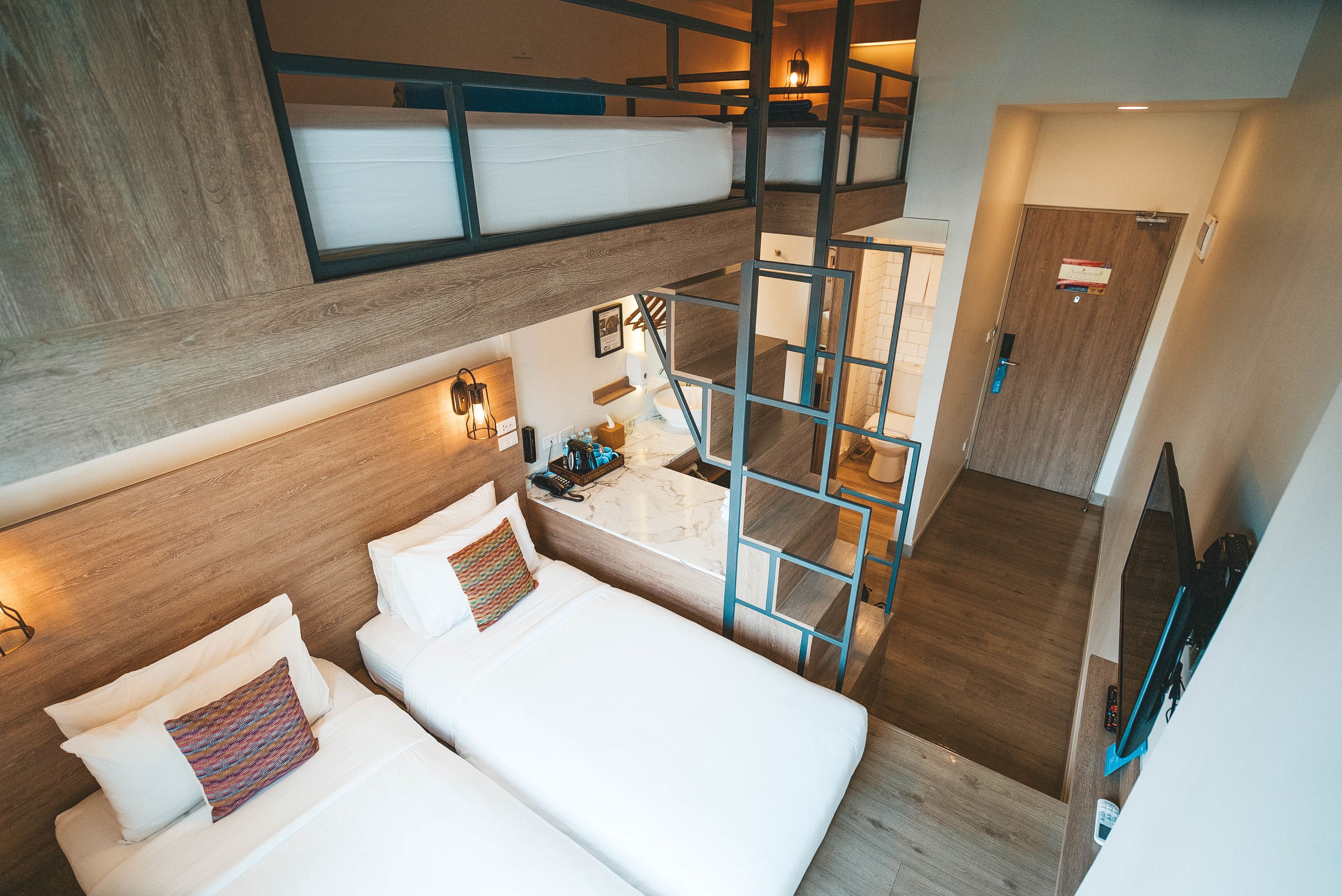 A modern hotel room with a loft bed, a small desk with a lamp, and a bathroom visible through a glass partition.