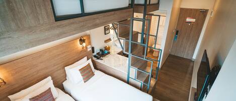 A modern hotel room with a loft bed, a small desk with a lamp, and a bathroom visible through a glass partition.