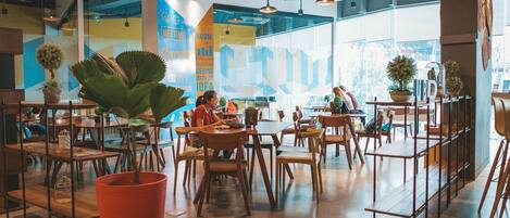 A modern cafe with wooden tables and chairs, potted plants, and large windows offering views of the street.