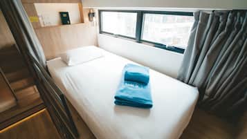 Ein Einzelbett mit weißer Bettwäsche und einem blauen Handtuch, ein Fenster mit Vorhängen, ein Holz-Kopfteil und eine kleine Ablagefläche mit einem Buch und einem Telefon.