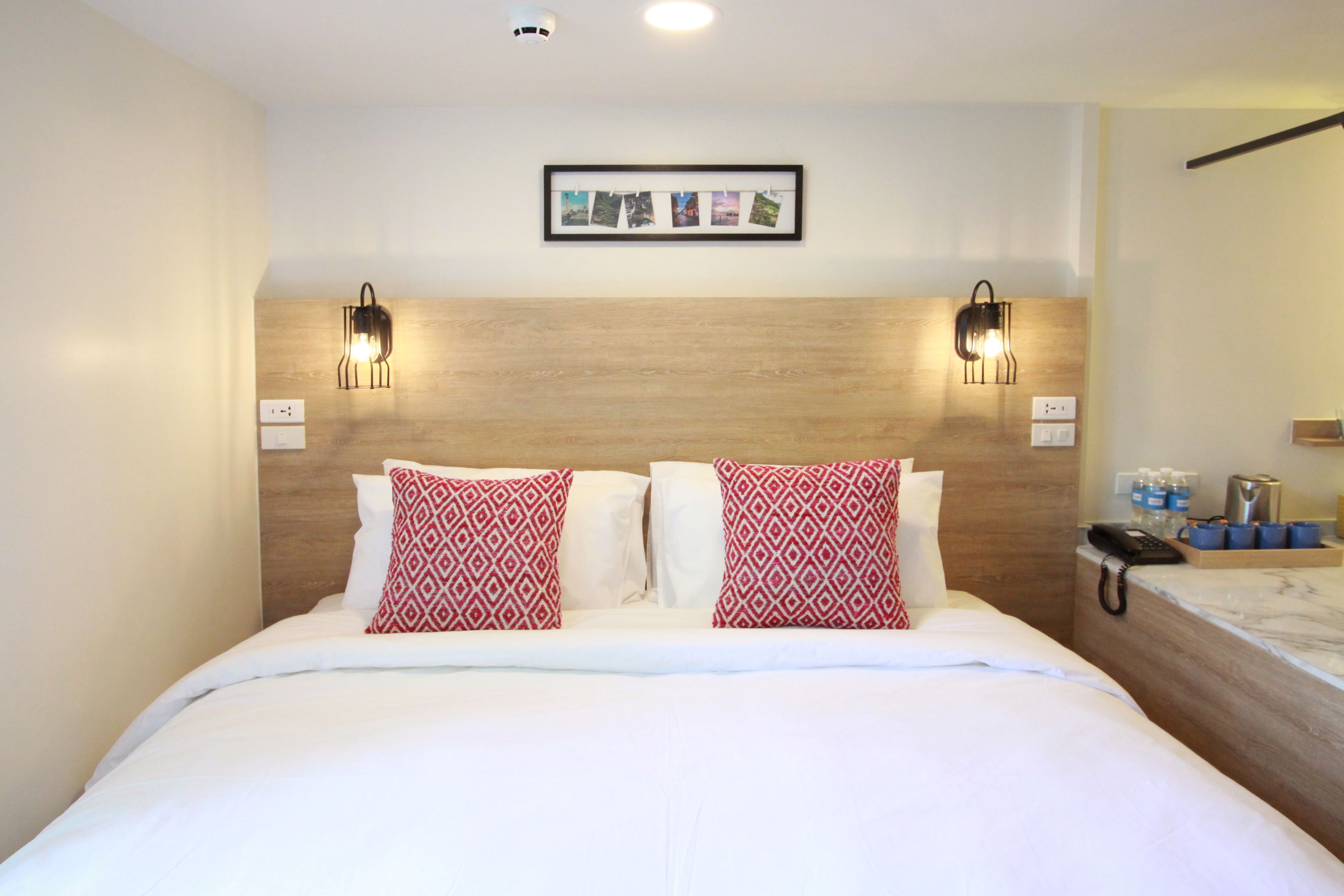 Une chambre d’hôtel avec une tête de lit en bois, du linge de lit blanc et deux coussins à motifs. Un cadre avec une photo est accroché au mur, et une table de chevet est posée à côté du lit, avec un téléphone et un porte-gobelet.
