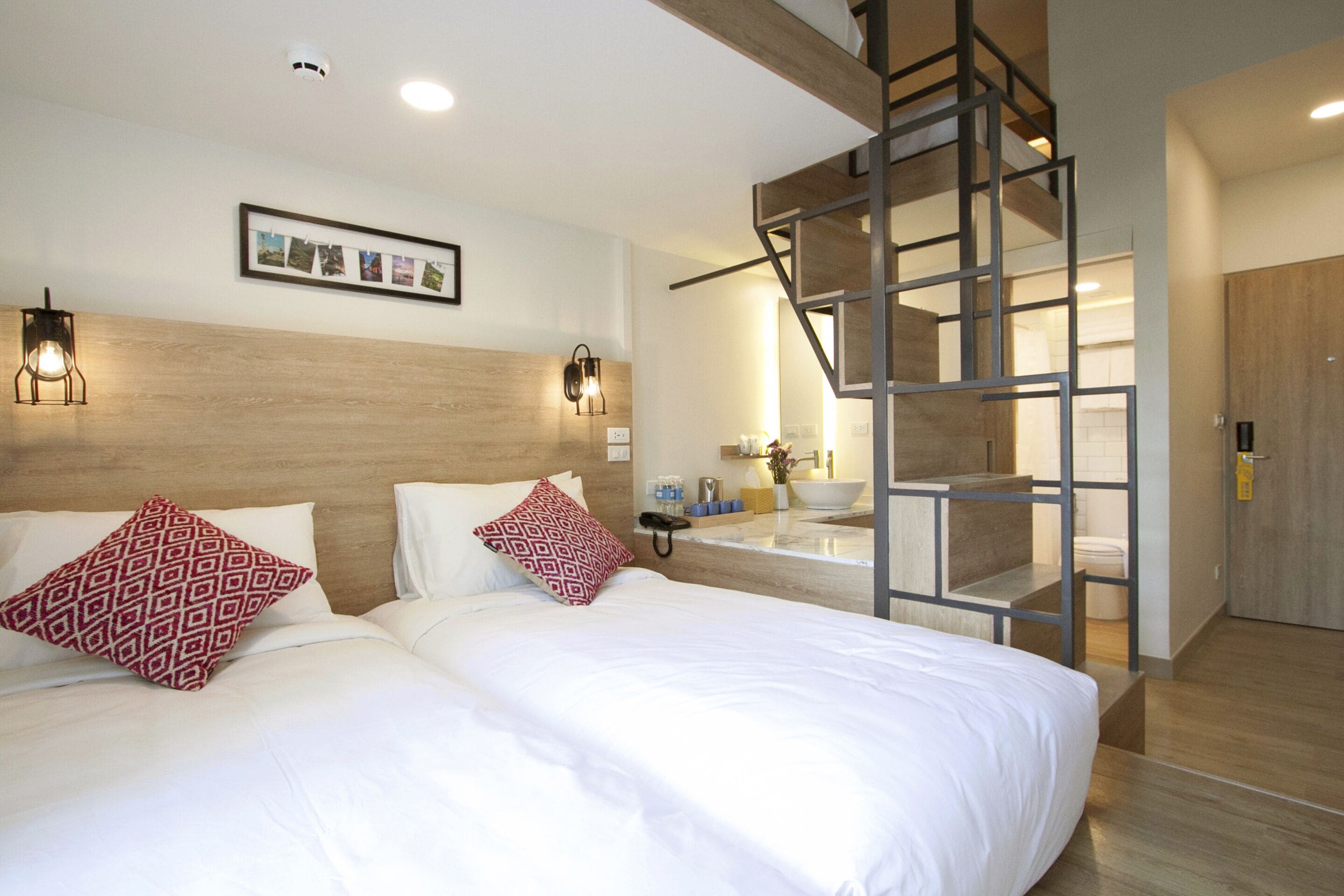 Ein modernes Hotelzimmer mit einem großen Bett, zwei gemusterten Kissen, einem hölzernen Kopfteil, einem gerahmten Foto, einer Treppe und einem Badezimmer mit Waschbecken und Spiegel.