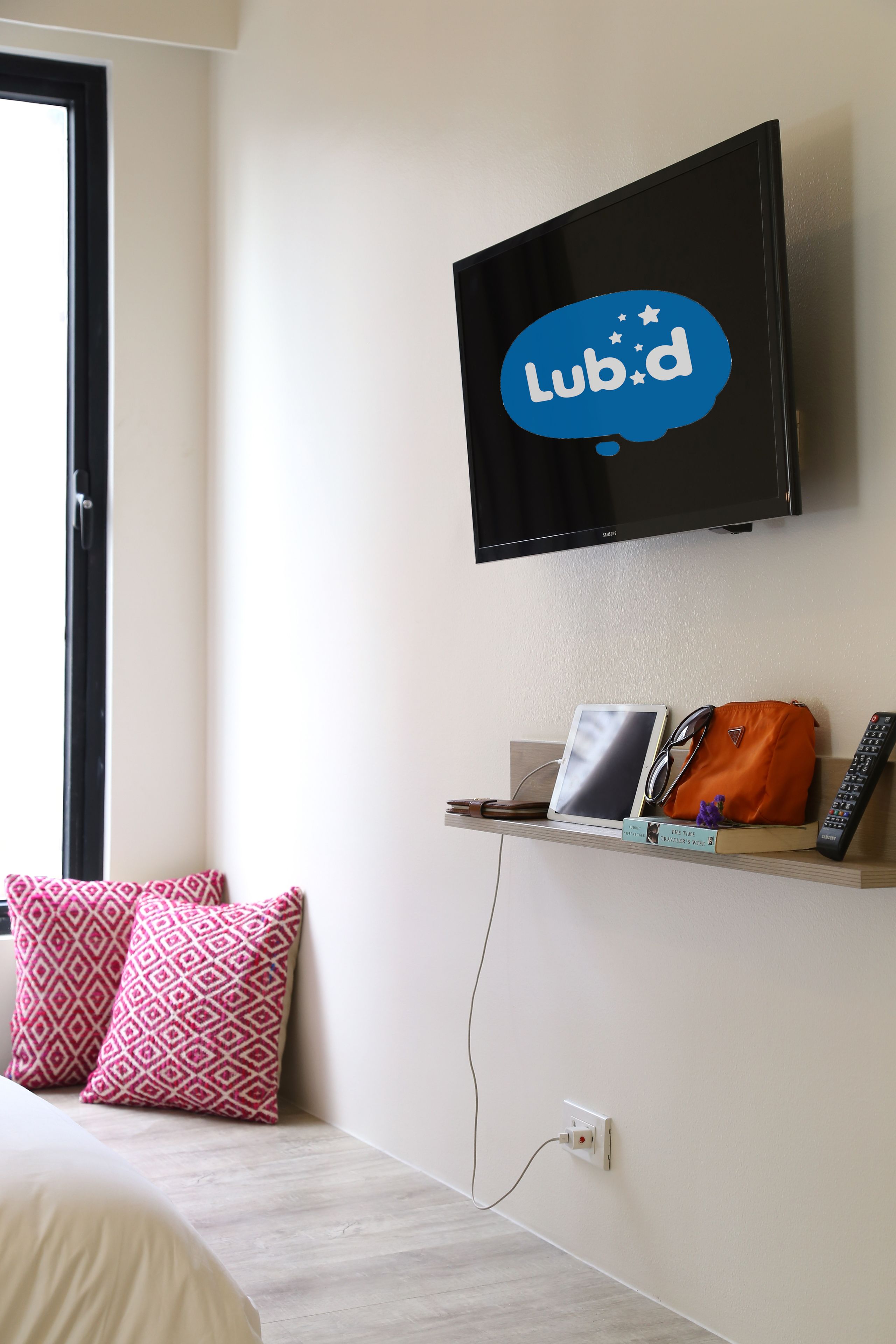 A hotel room with a bed, pillows, a TV mounted on the wall, a shelf with a remote, a tablet, and a handbag.