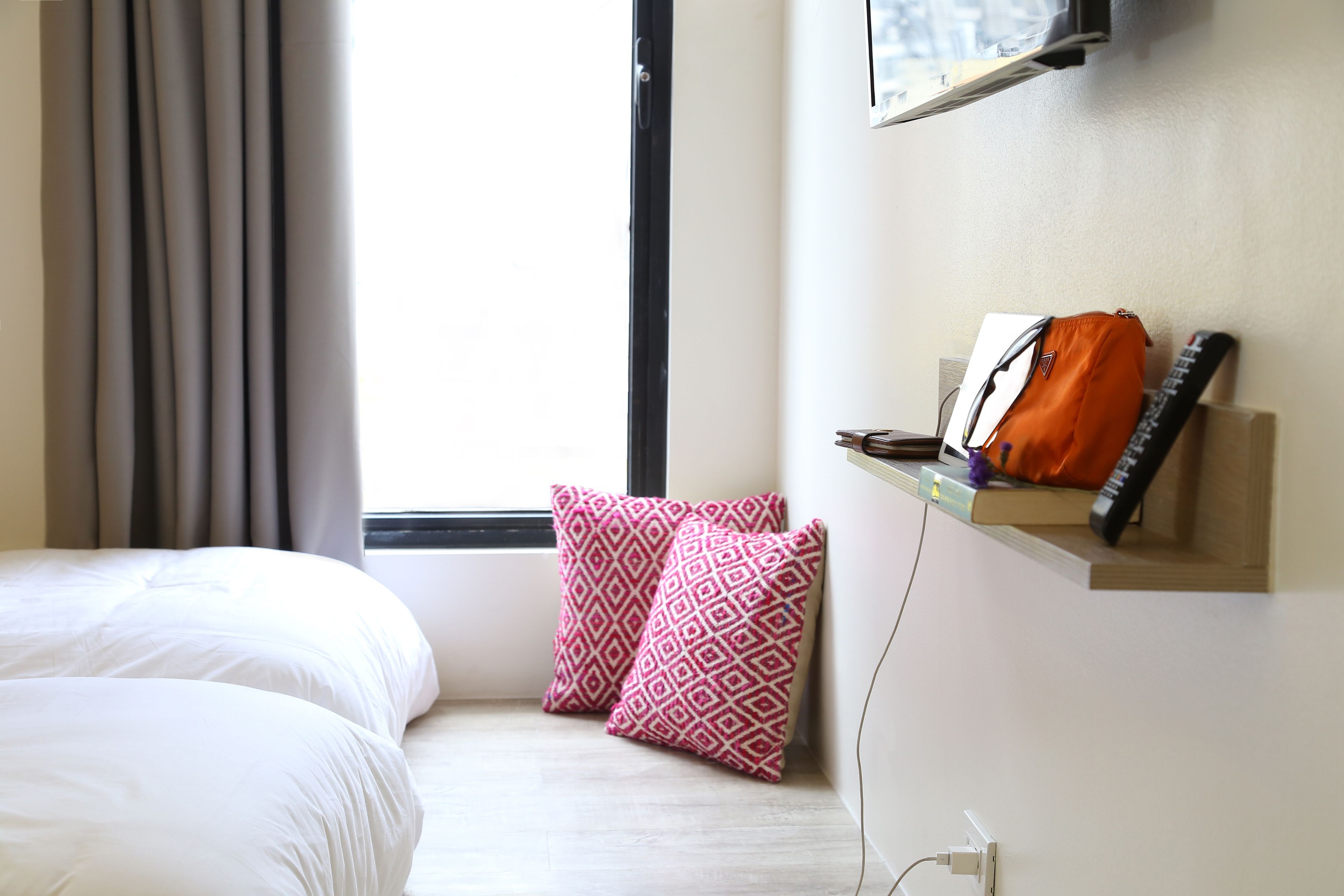 A hotel room with a bed, two patterned pillows, a shelf with a bag and remote, and a window with curtains.