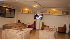 Een ruime woonkamer met een hoekbank, een rode fauteuil en een bruine fauteuil. Een flatscreen televisie is aan de muur bevestigd, en daarboven staan schappen met decoratieve objecten.