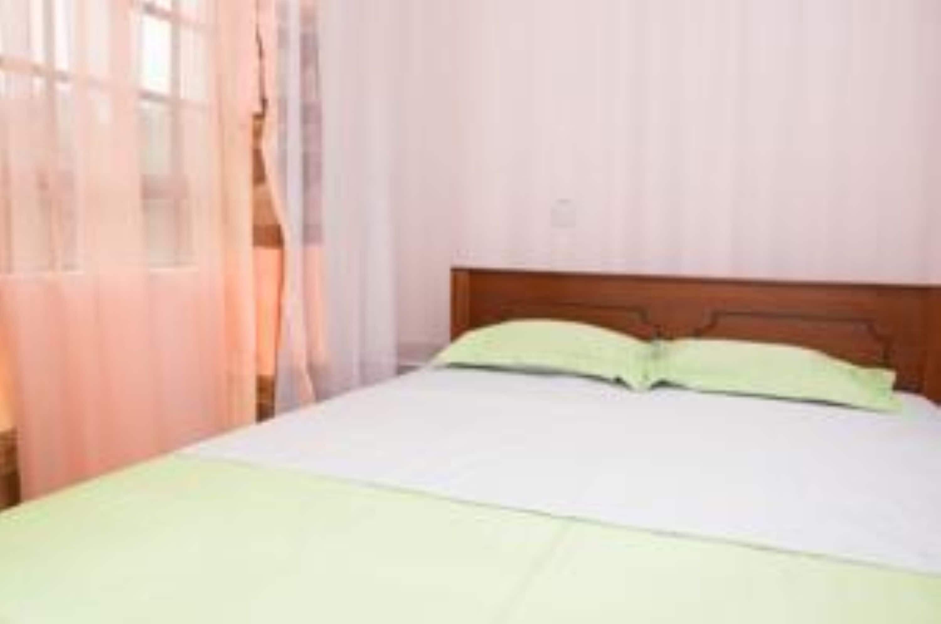 Una cama individual con cabecera de madera, almohada verde y cubrecama blanco en una habitación con ventana cubierta por cortinas traslúcidas.