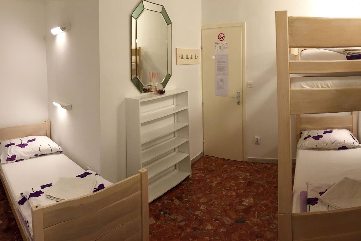 A dormitory room with two single beds, a bunk bed, a mirror, a white wardrobe, and a bulletin board.