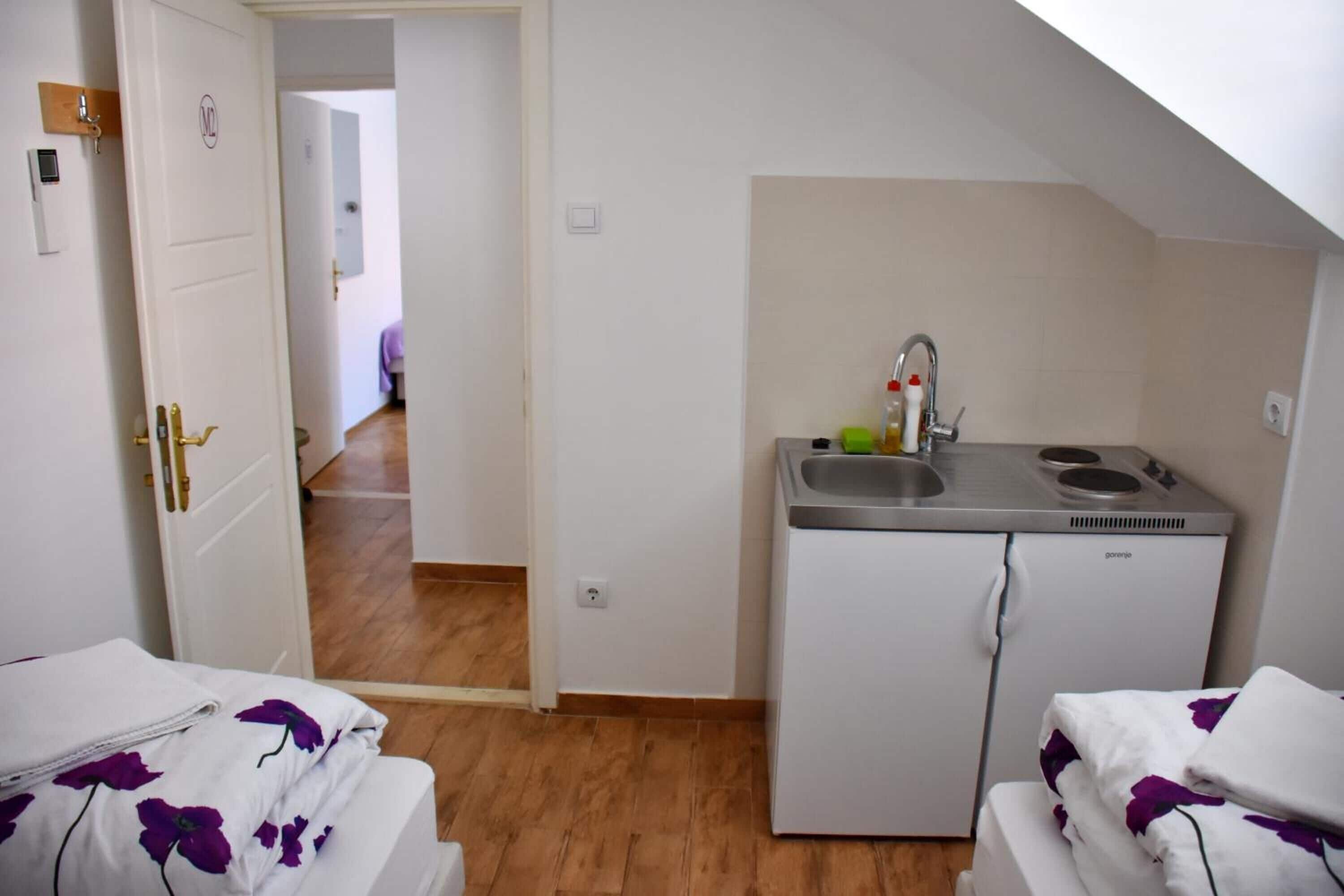 Cocina compacta con lavamanos y refrigerador, cama con ropa de cama floral, y puerta que da a otra habitación.
