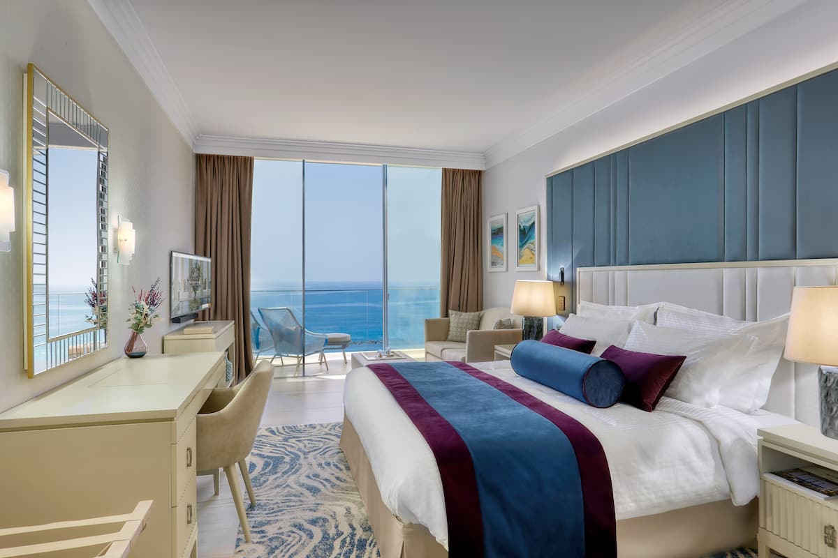 A hotel room with a large bed, a desk, a chair, a nightstand, and a view of the ocean.
