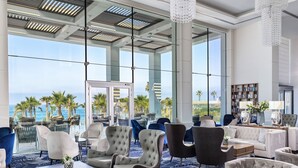 A spacious lounge area with comfortable seating, large windows offering a view of the ocean, and a modern ceiling design. - Amavi, MadeForTwo Hotels - Paphos (Paphos)